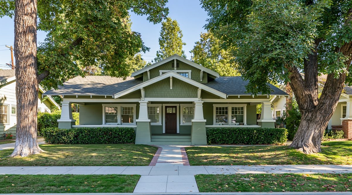 Front of a sage-green Craftsman bungalow with a covered porch and shade trees